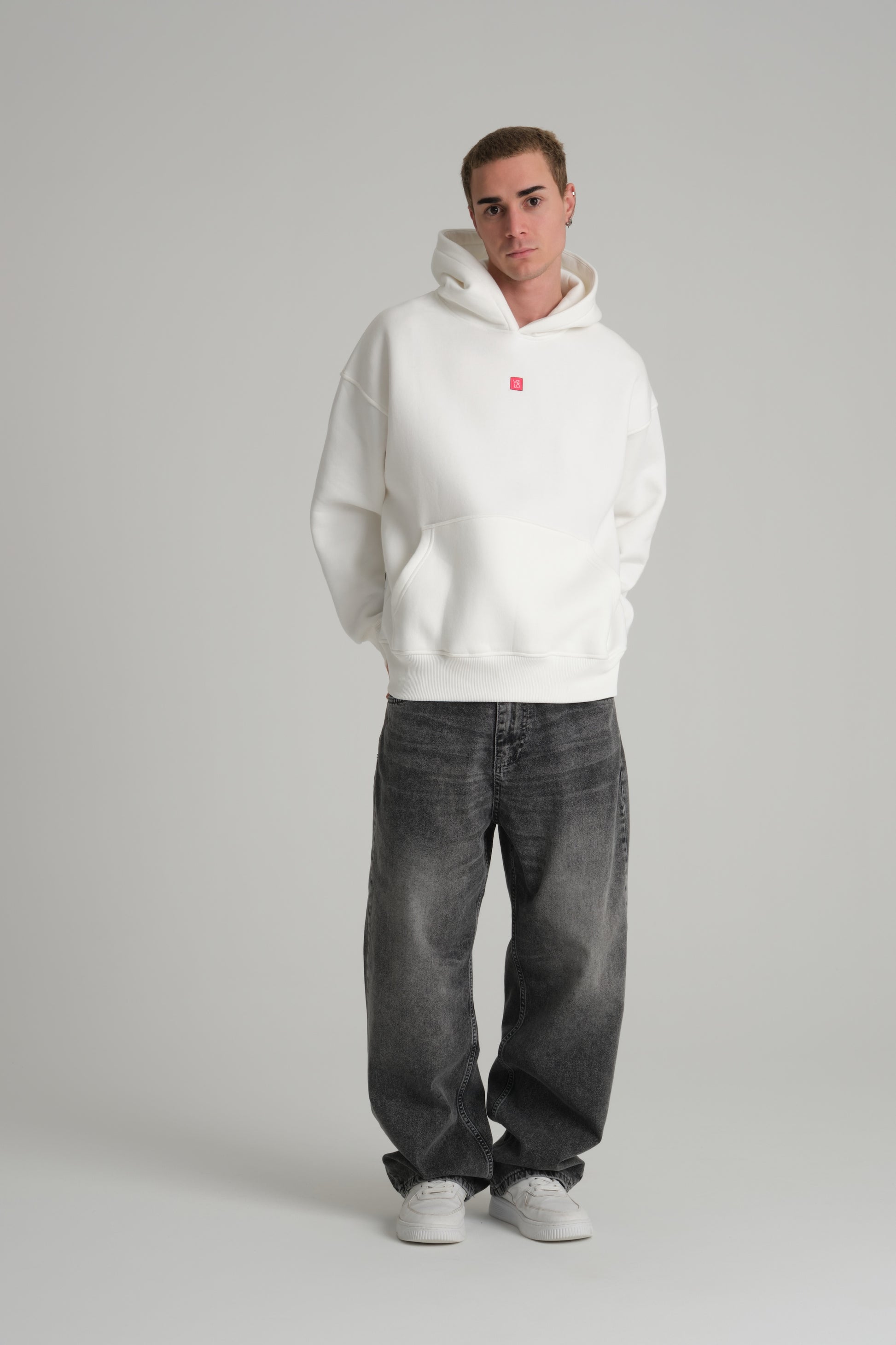 Person wearing a white hoodie with a red logo and dark jeans on a gray background