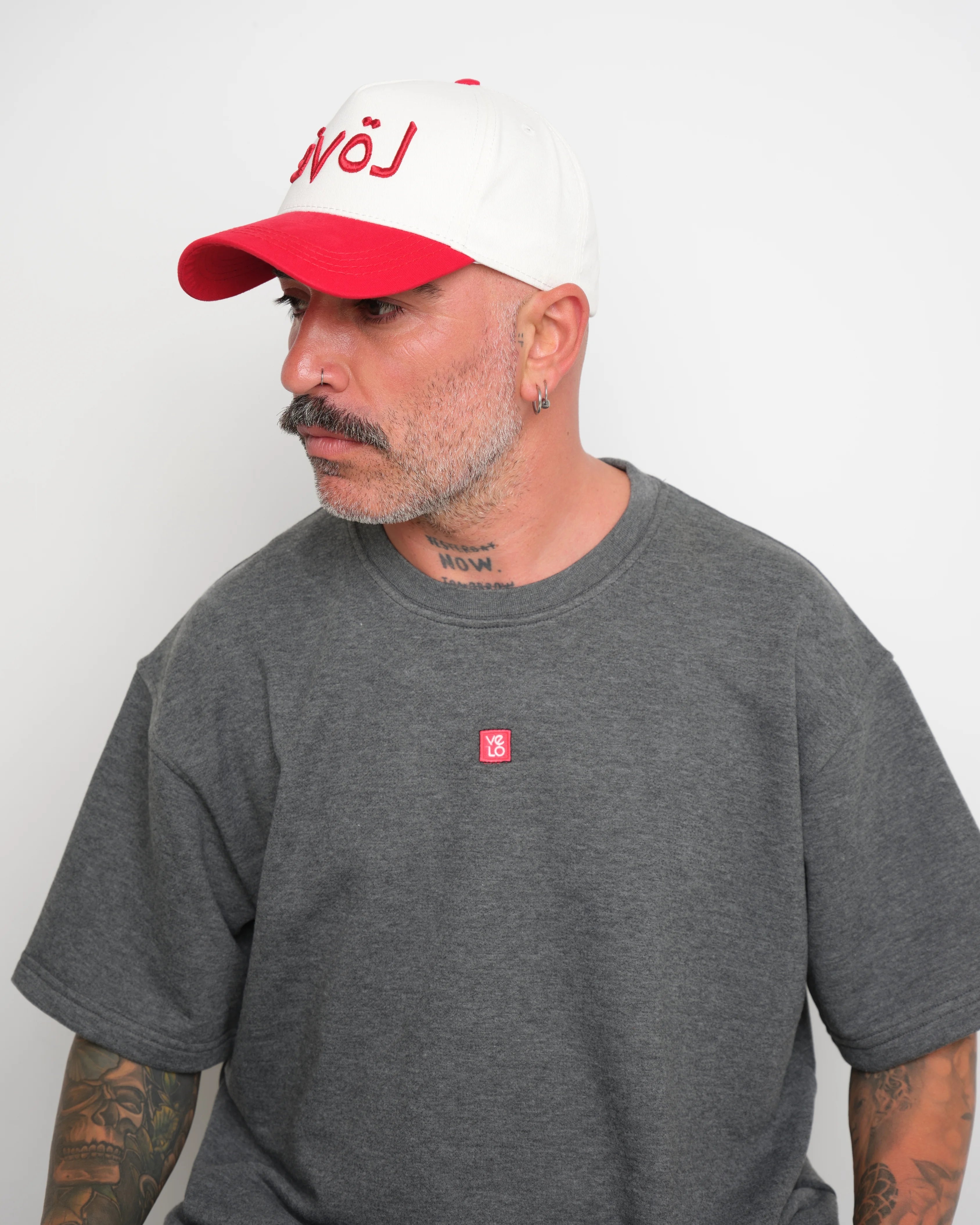 Man wearing a gray t-shirt with 'velo' geometric stitched and white baseball cap with red trim and Arabic red 'love' letter on a white background
