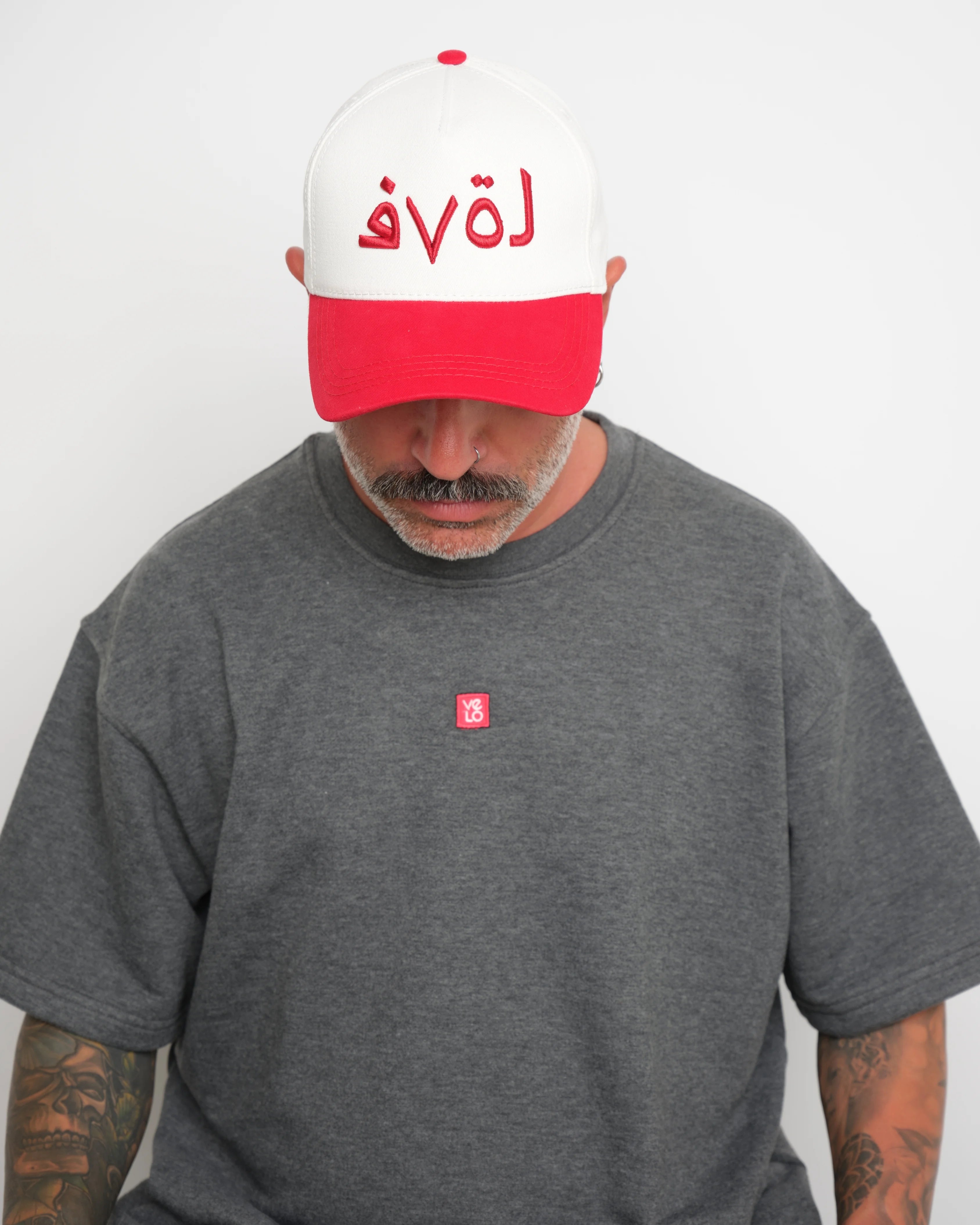 Man wearing a red and white cap with red brim and Arabic 'love'  text and a gray t-shirt with a small VELO logo.