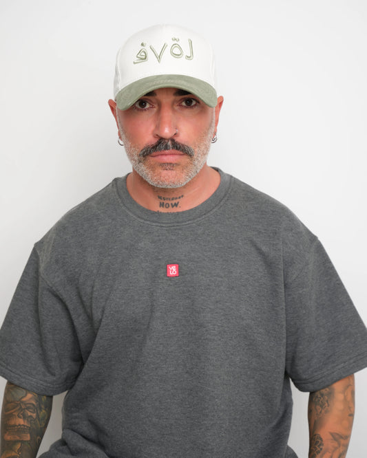 Man wearing a gray t-shirt with a VELO logo and a white cap with sage green brim and Arabic 'love'  text on a plain background