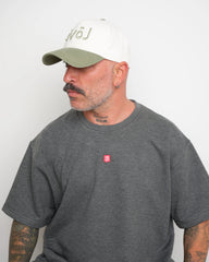 Side look of man wearing a gray t-shirt with a VELO logo and a white cap with sage green brim and Arabic 'love'  text on a plain background