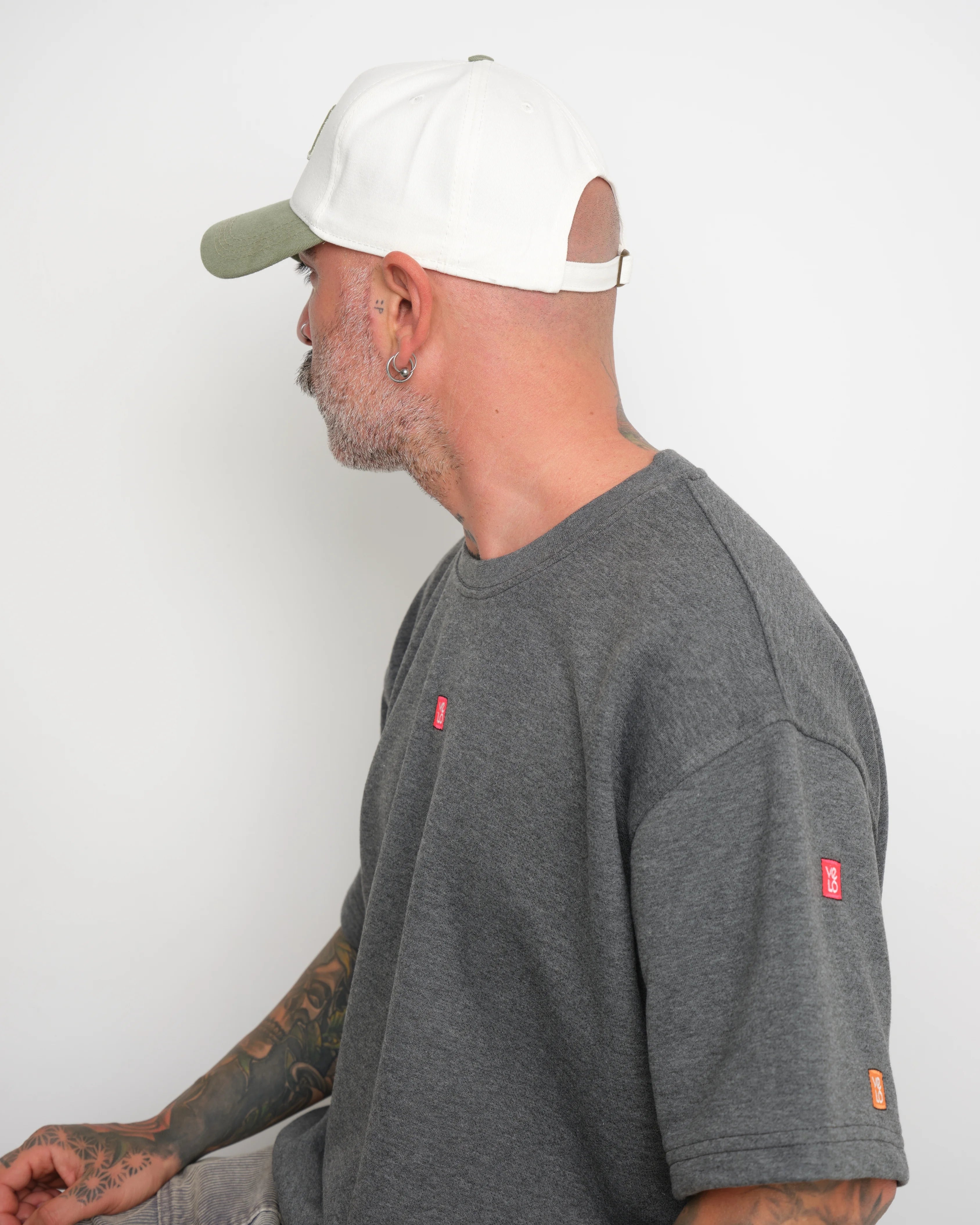 Man wearing a gray t-shirt with small logos on a white background and shows adjustable flip tag of the cap