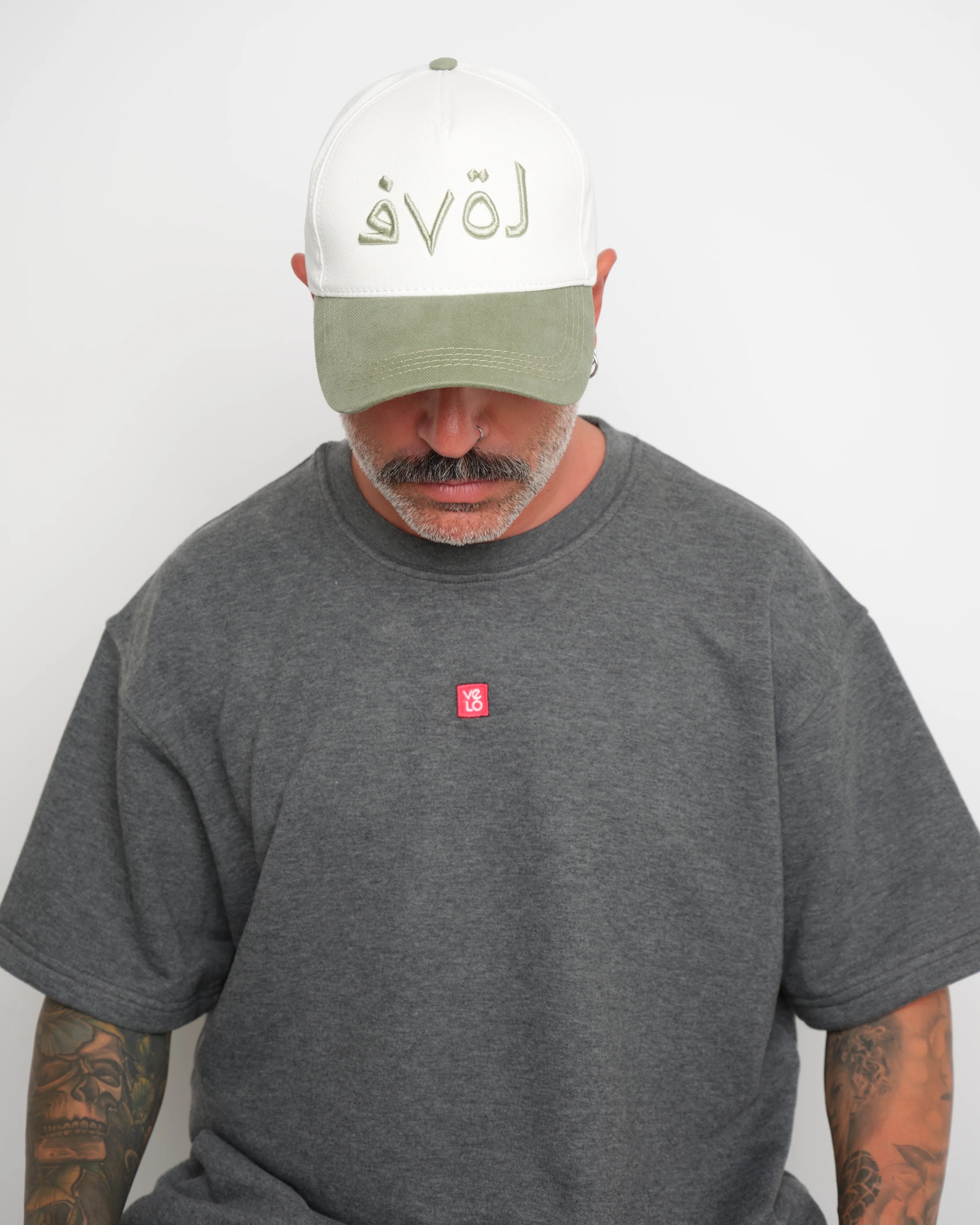 Man wearing a gray t-shirt with a red 'VELO' logo and a white cap with safe green brim and Arabic 'LOVE' text.