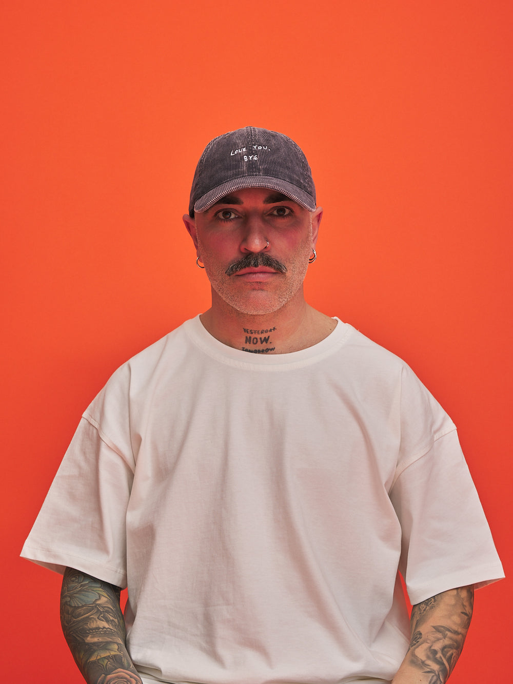Man wearing a charcoal grey corduroy cap and white t-shirt against an orange background