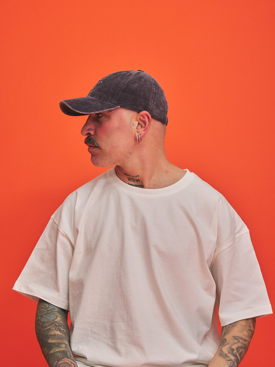 Person wearing a charcoal grey corduroy cap and white t-shirt against an orange background