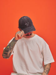 Person wearing a charcoal grey corduroy cap and white t-shirt against an orange background