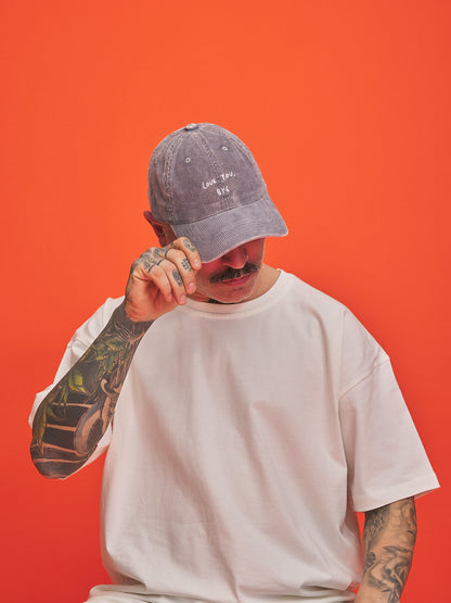 Person wearing a stone grey corduroy cap and white t-shirt against an orange background