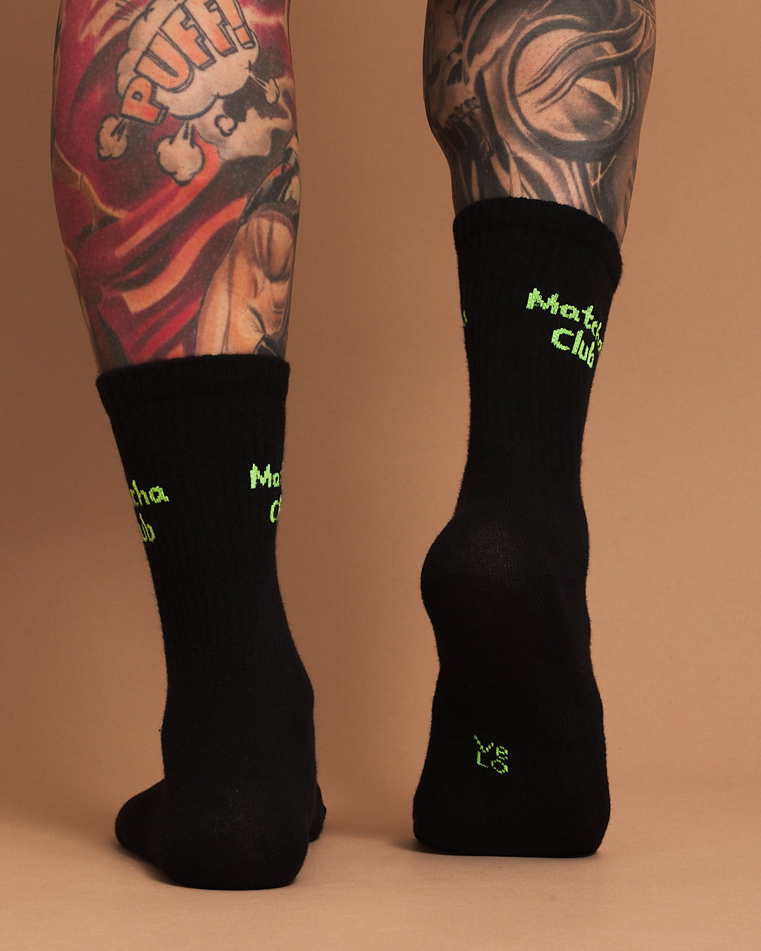 Black socks with green text worn by a person with tattoos on a orange background