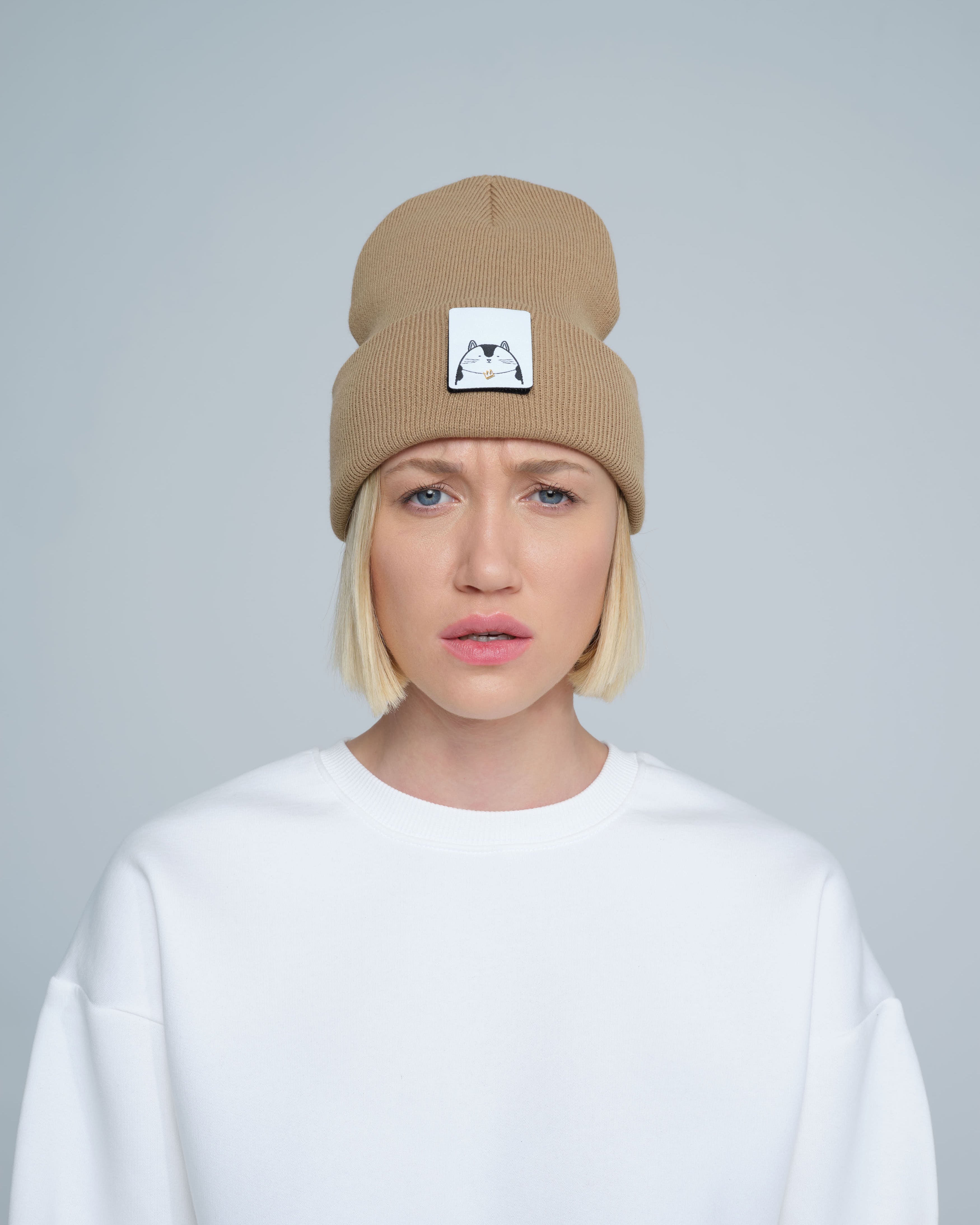 A person wearing mink brown colour beanie with cat illustration patch on the front