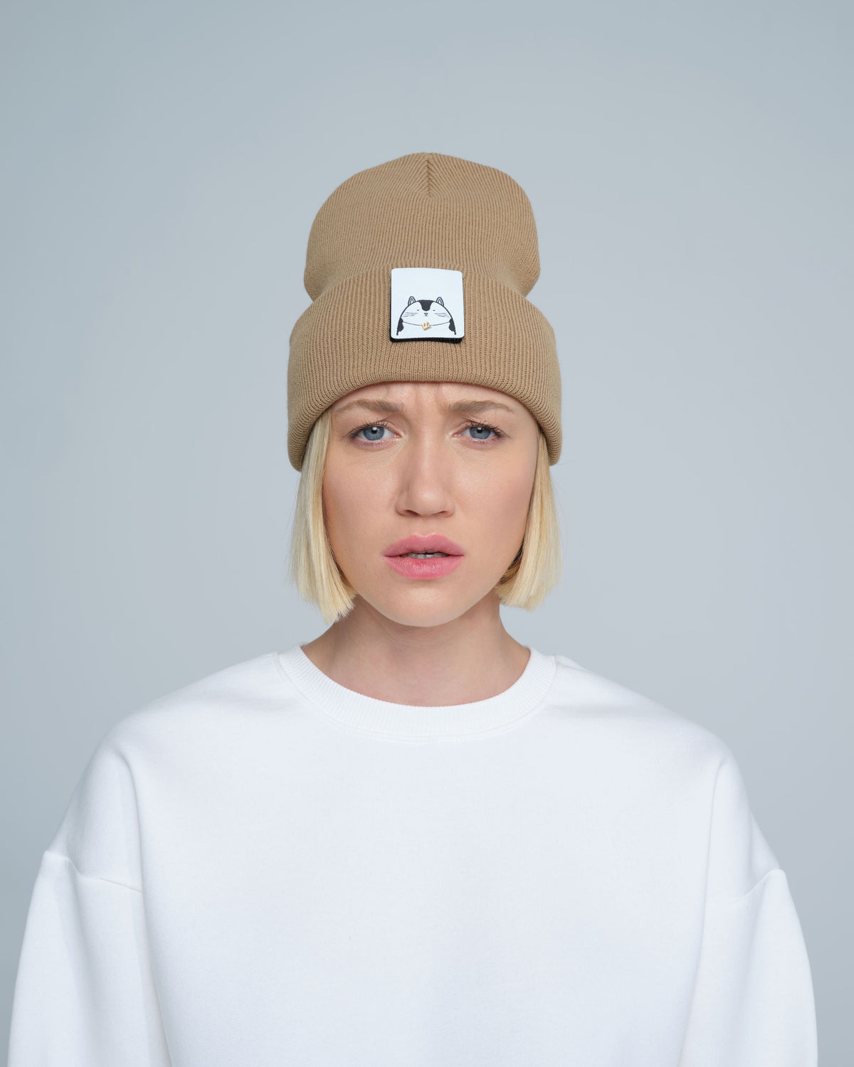 A person wearing mink brown colour beanie with cat illustration patch on the front