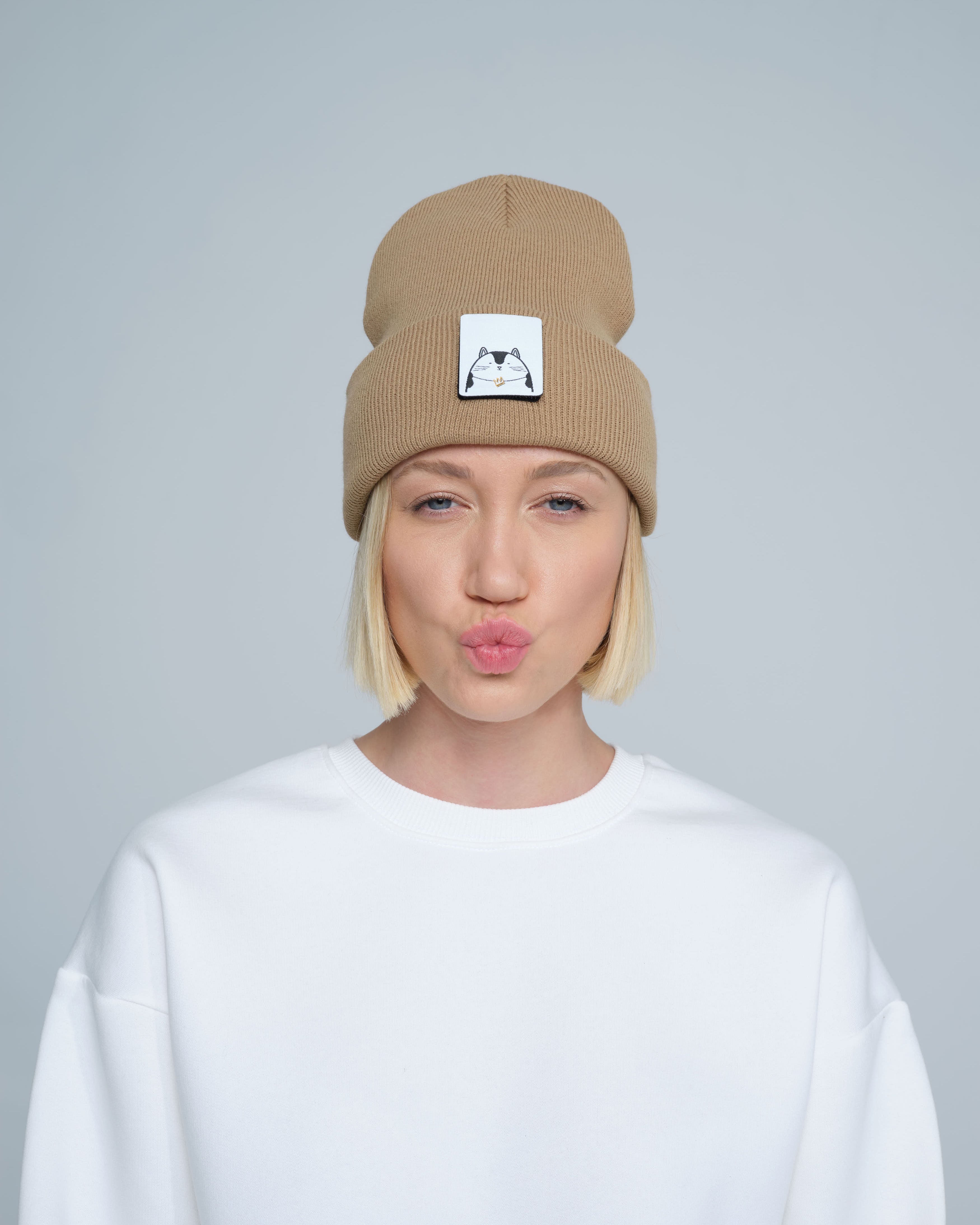 A person wearing mink brown colour beanie with cat illustration patch on the front on a gray background