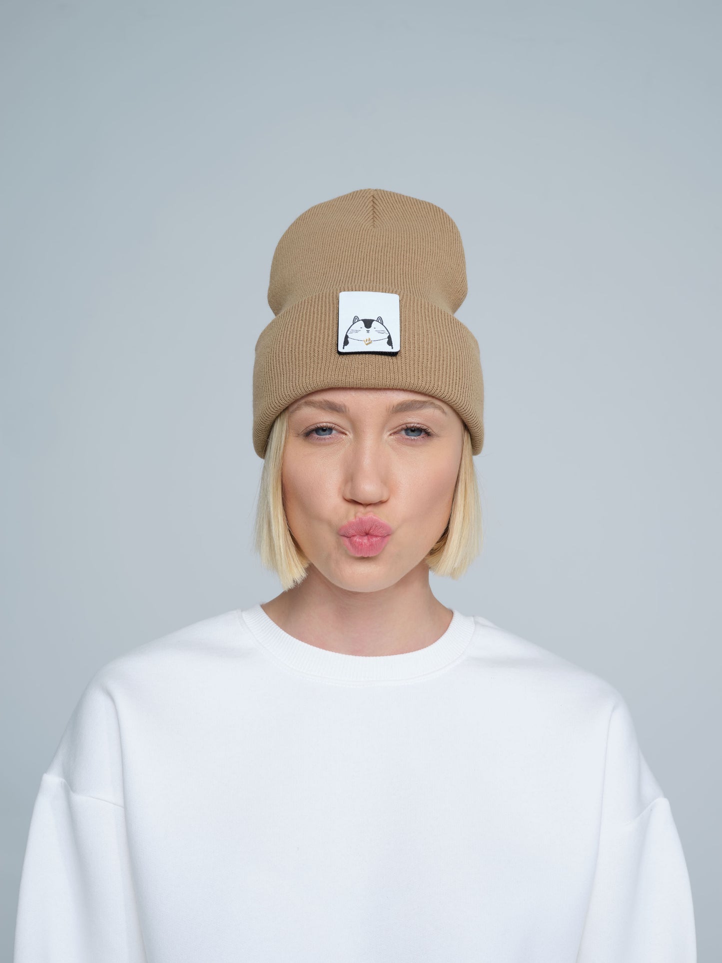 A person wearing mink brown colour beanie with cat illustration patch on the front on a gray background