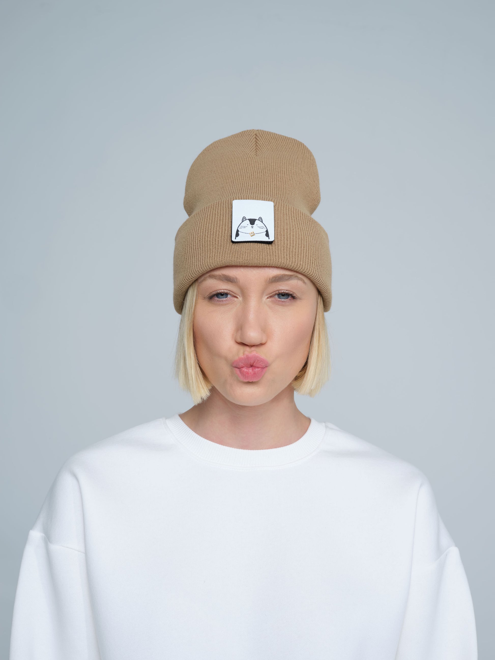 A person wearing mink brown colour beanie with cat illustration patch on the front on a gray background