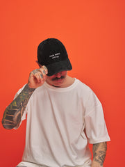 Person wearing a black cap with text, white t-shirt, and tattoos on an orange background