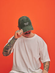 Person wearing a green cap with text, white t-shirt, and tattoos on an orange background