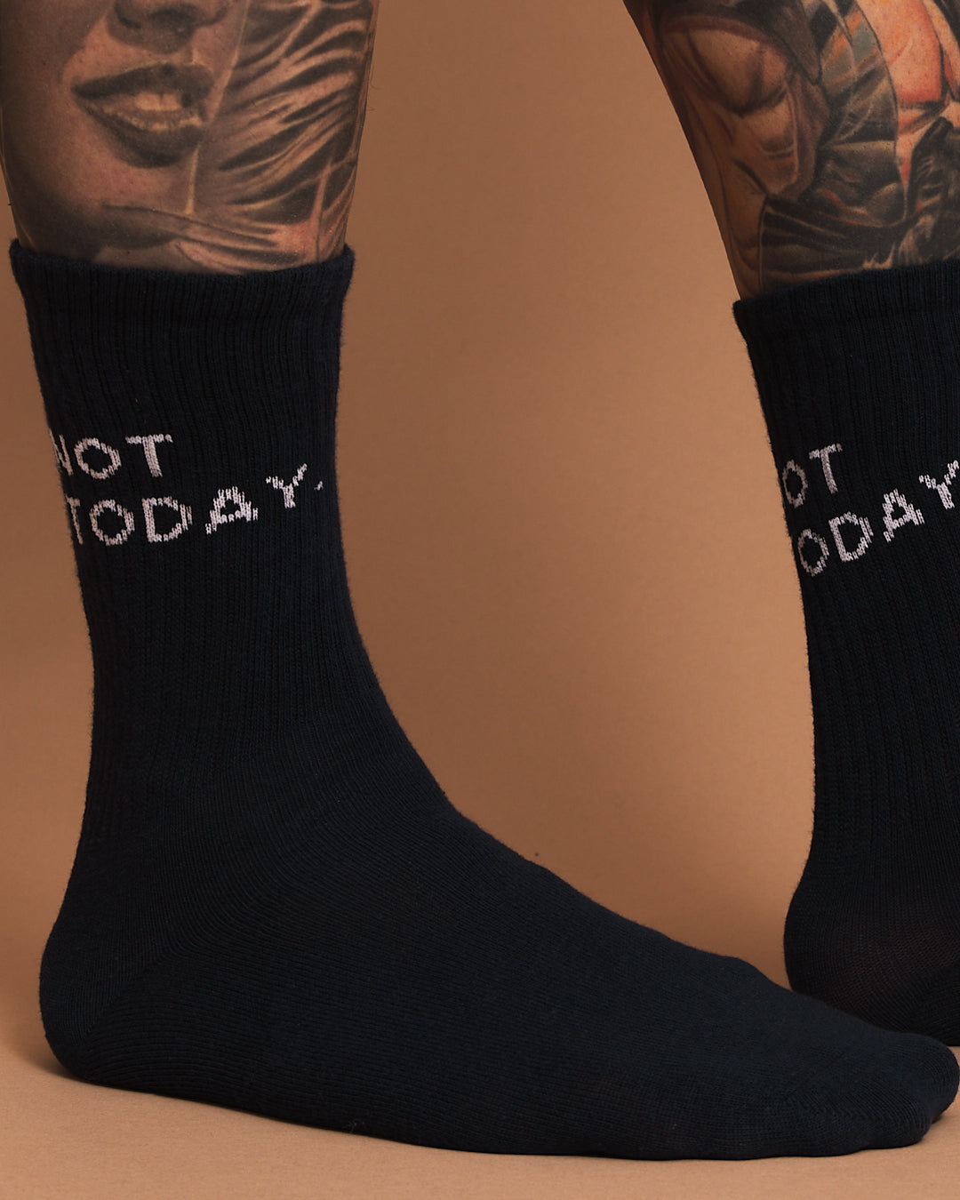Side look of navy socks with 'Not Today' text worn by a person with tattoos on a brown background