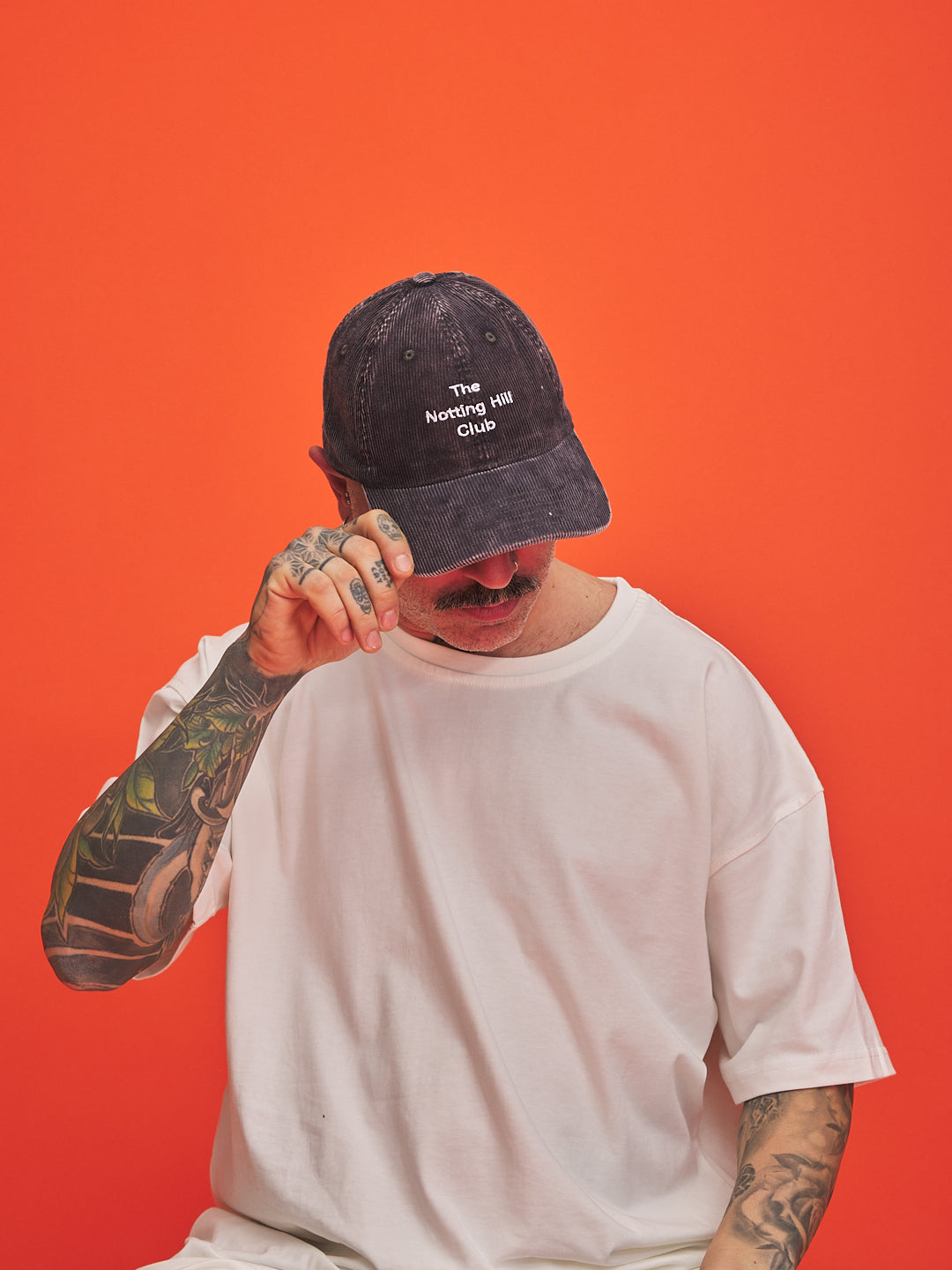 Person wearing a grey cap with text, white t-shirt, and tattoos on an orange background