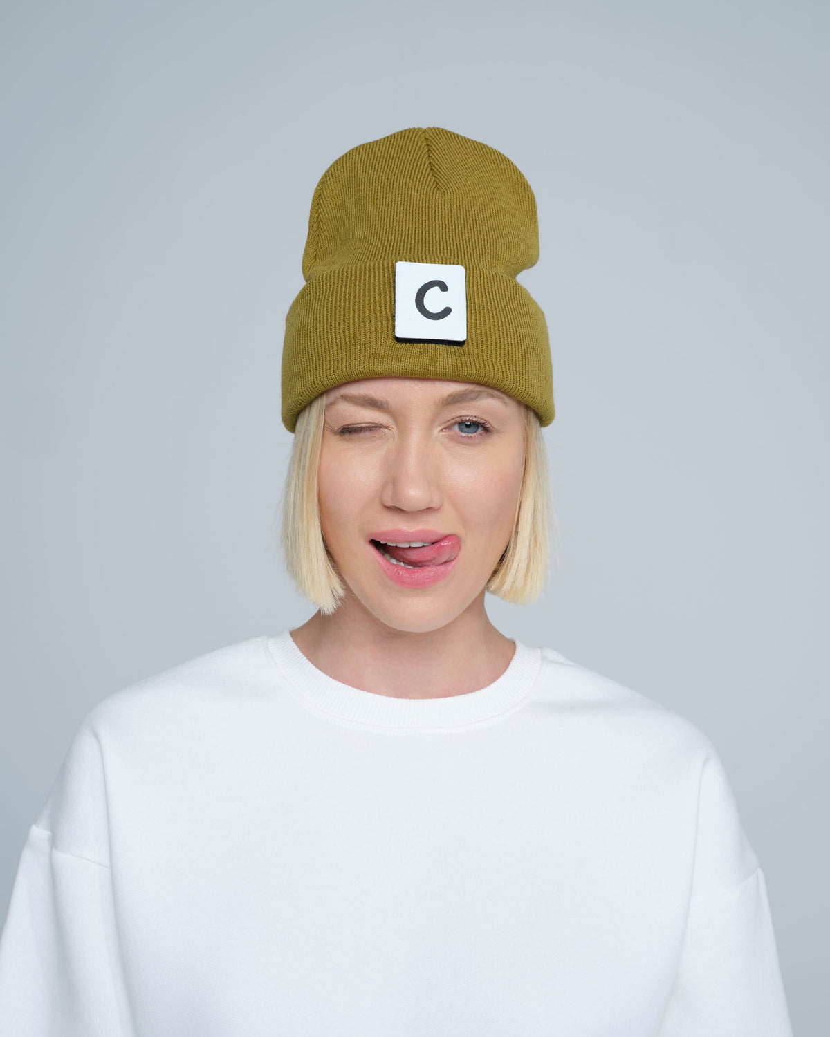 A person wearing olive beanie with 'C' letter patch attached on front on a light gray background