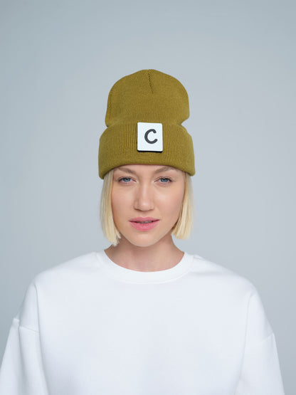 A person wearing olive beanie with 'C' letter patch attached on front on a light gray background