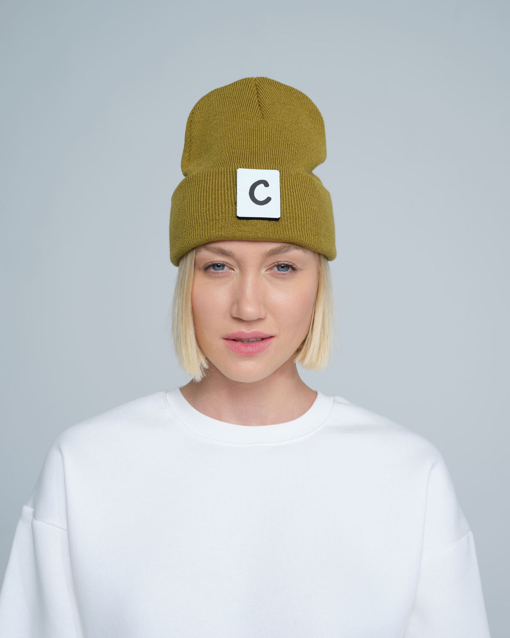 A person wearing olive beanie with 'C' letter patch attached on front on a light gray background