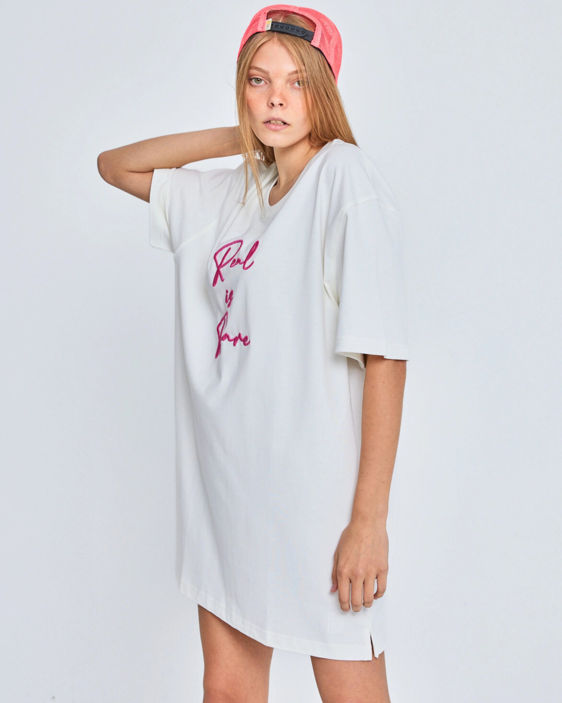 Person wearing a white oversized t-shirt with pink text and a red cap on a light gray background