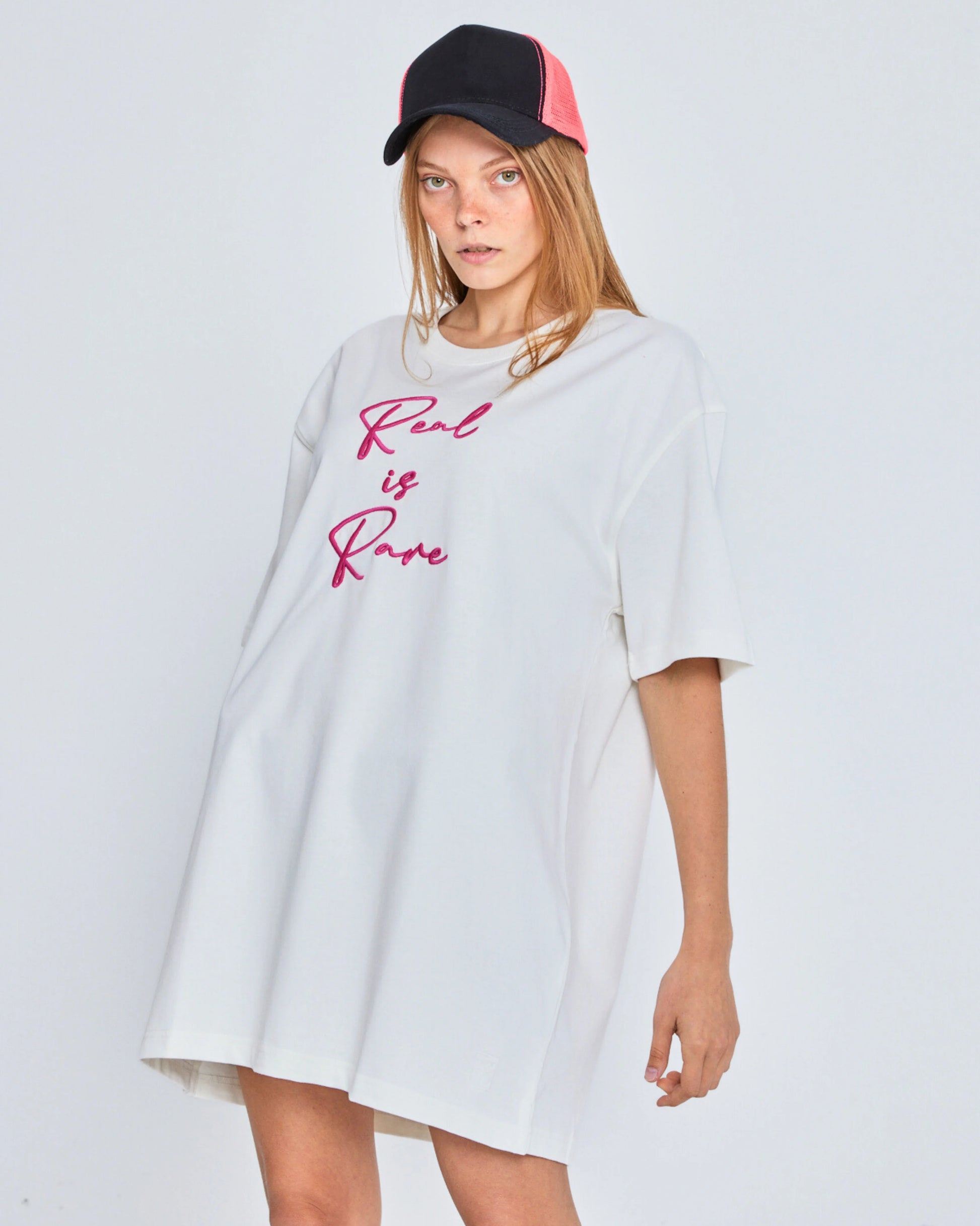 Person wearing a white oversized t-shirt with 'Real is Rare' text and a black and black cap on a light gray background