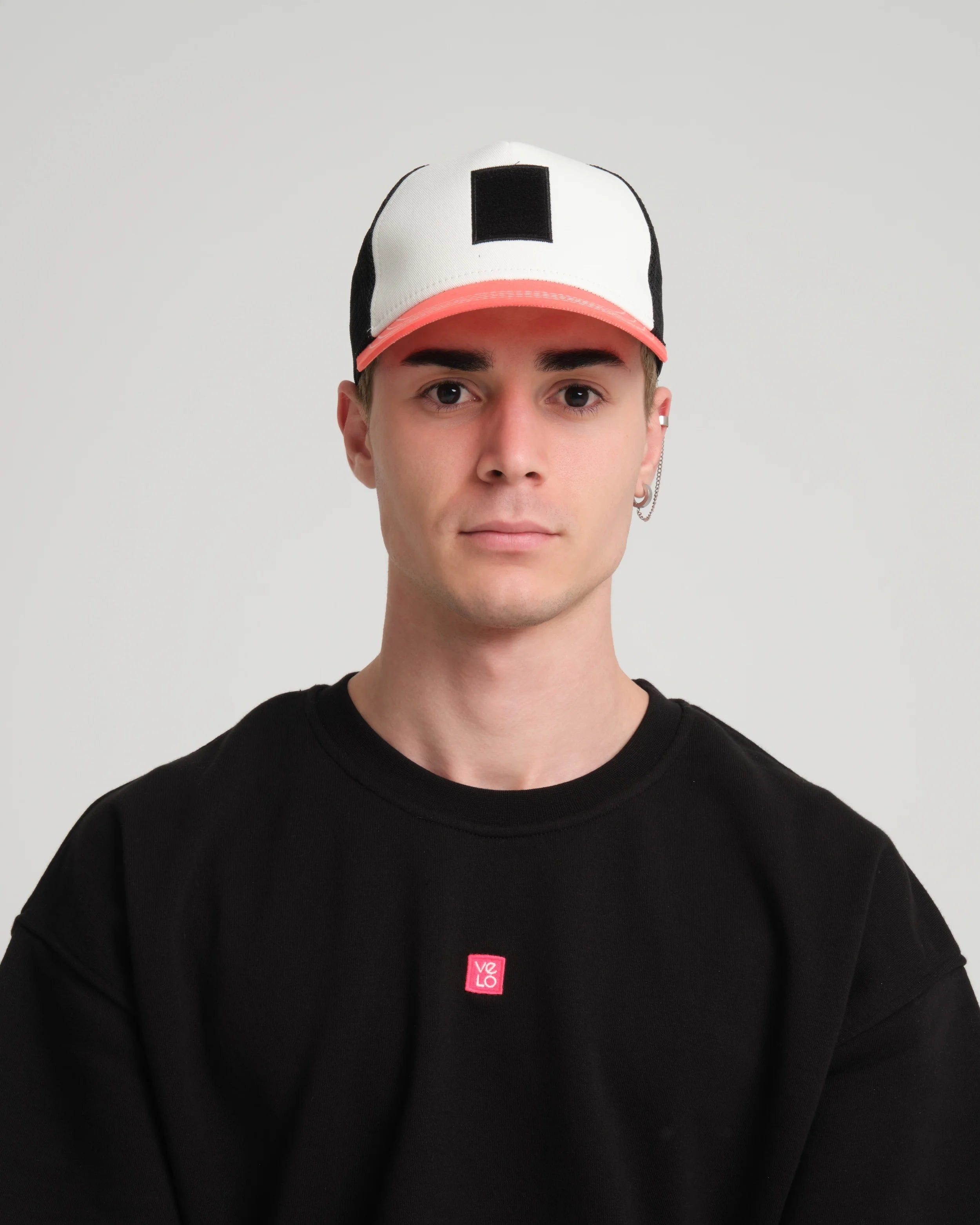 Person wearing a black t-shirt with a small red logo and a white and pink cap on a gray background