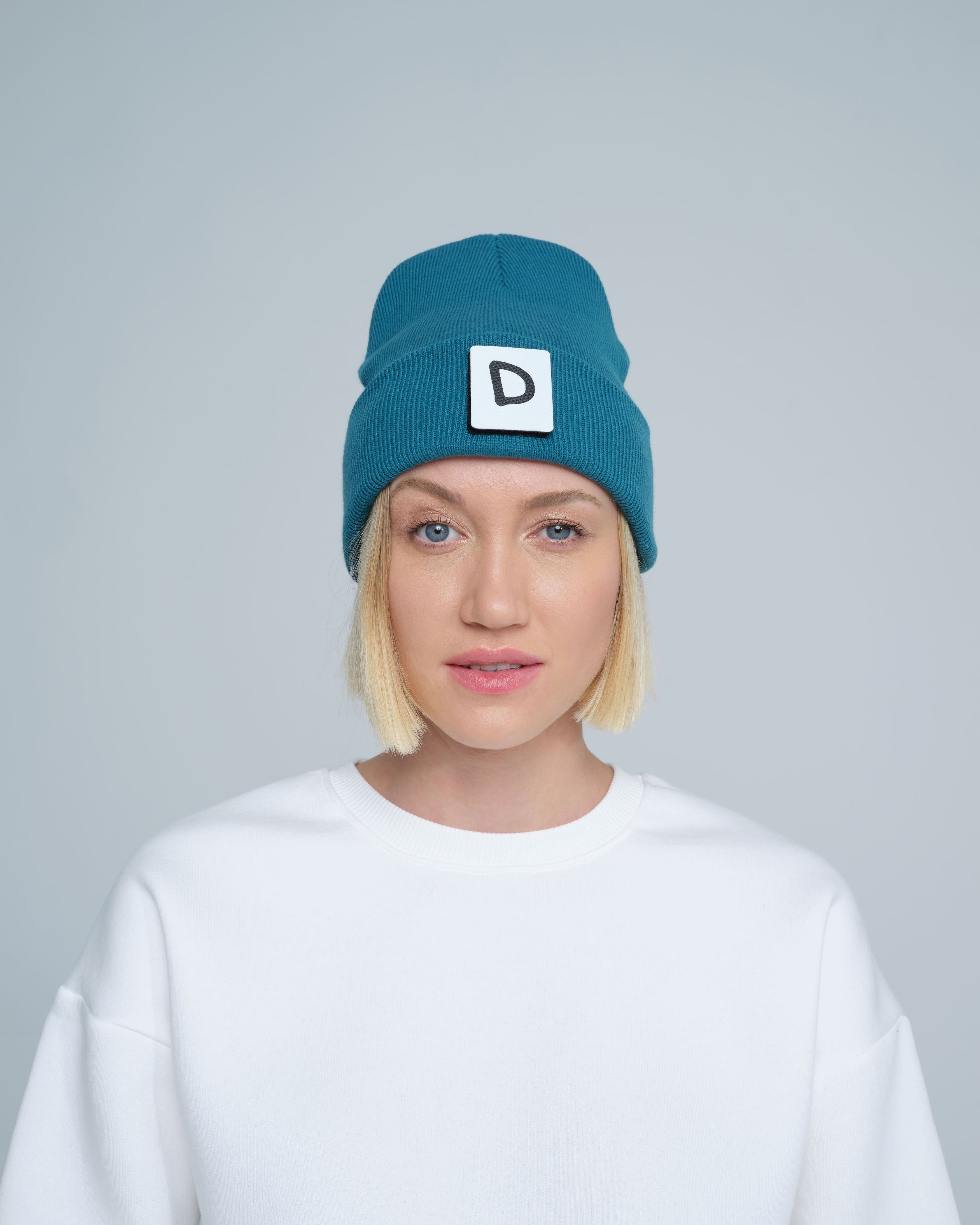 A person wearing teal beanie with 'D' letter patch attached on the front on a light gray background