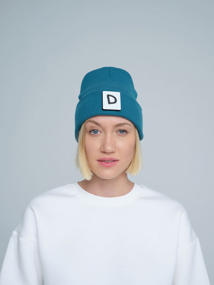 A person wearing teal beanie with 'D' letter patch attached on the front on a light gray background