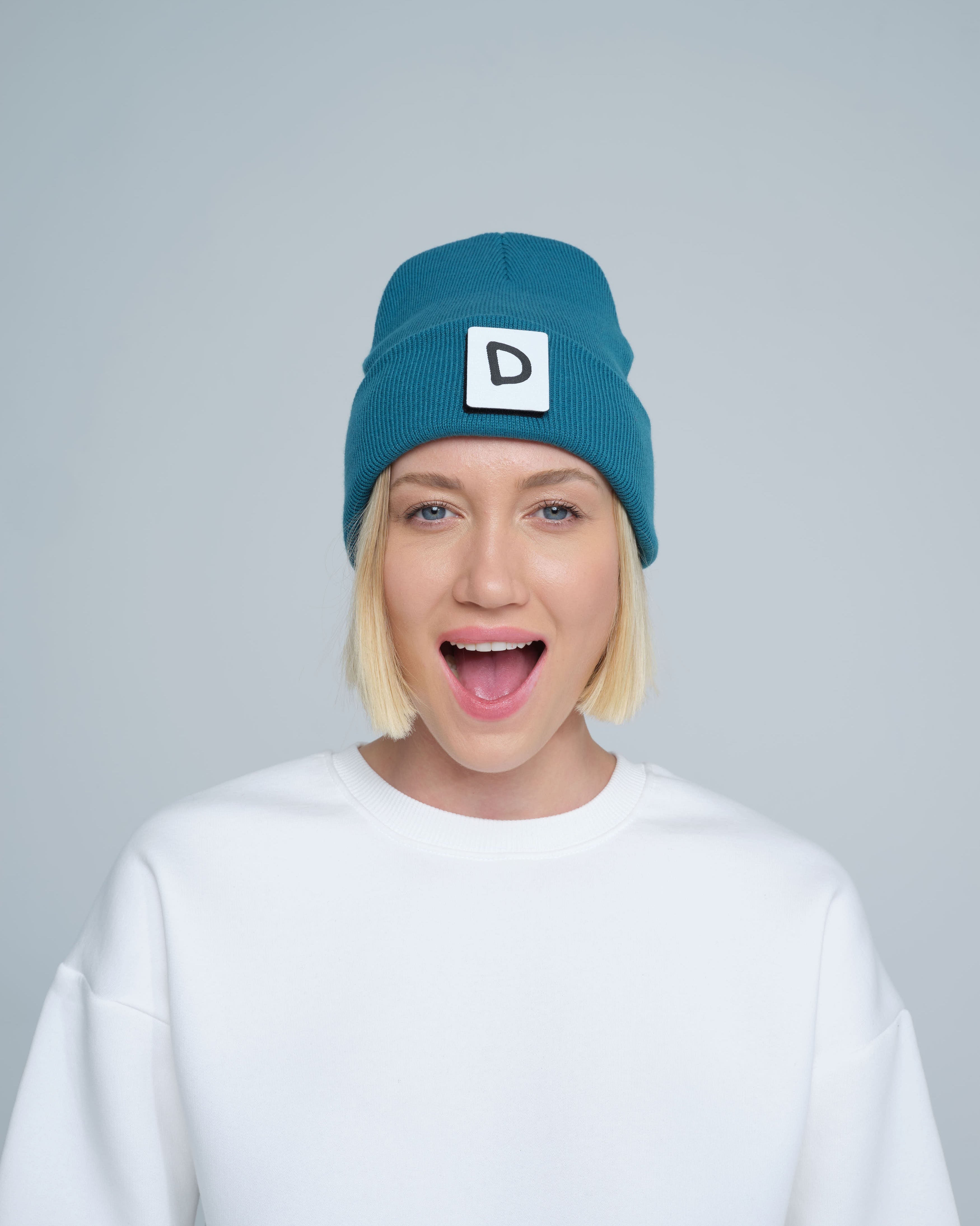 A person wearing teal beanie with 'D' letter patch attached on the front on a light gray background