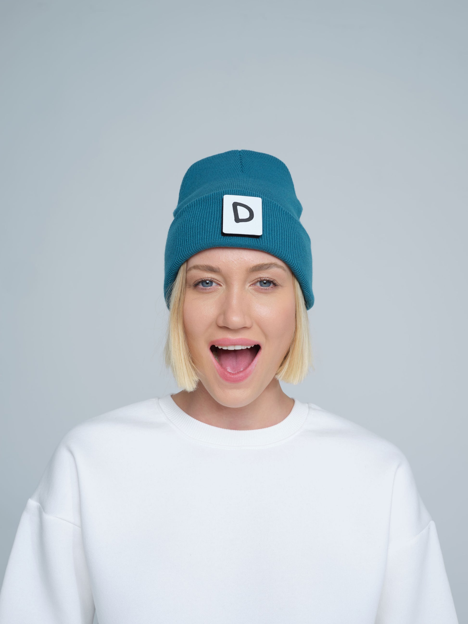A person wearing teal beanie with 'D' letter patch attached on the front on a light gray background