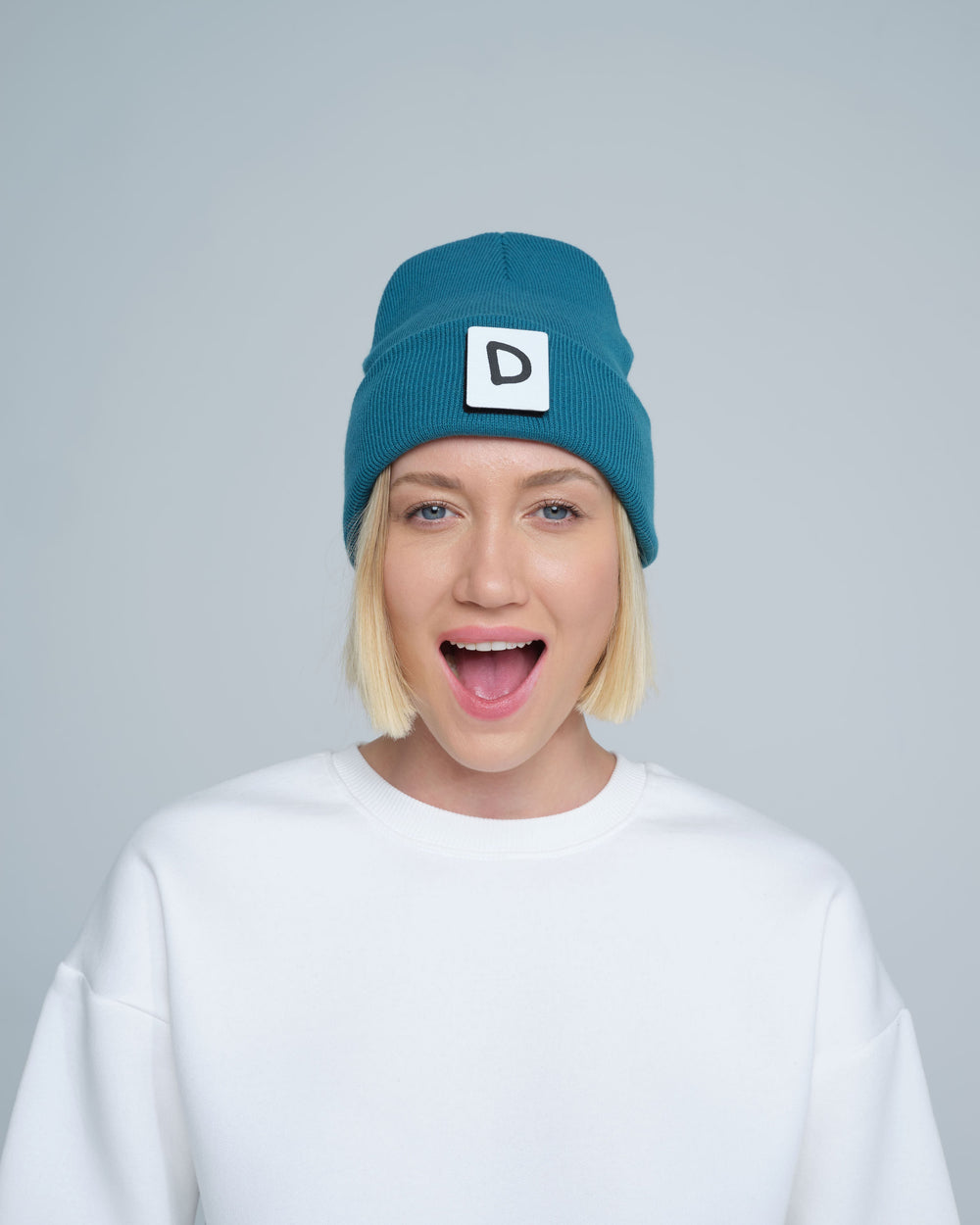 A person wearing teal beanie with 'D' letter patch attached on the front on a light gray background