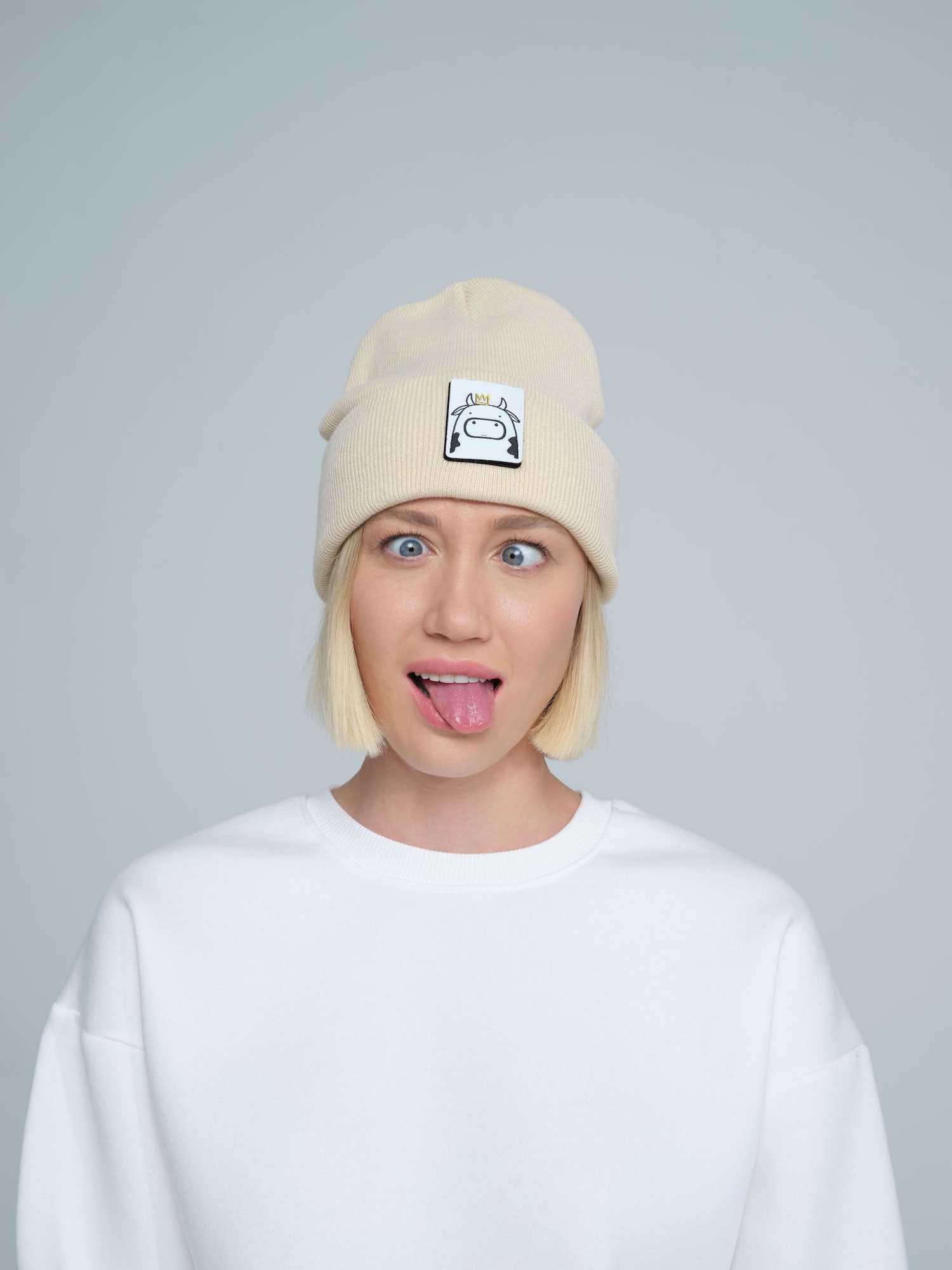A person wearing a beige beanie hat with a 'cow' design on the cuff, paired with a white t-shirt.
