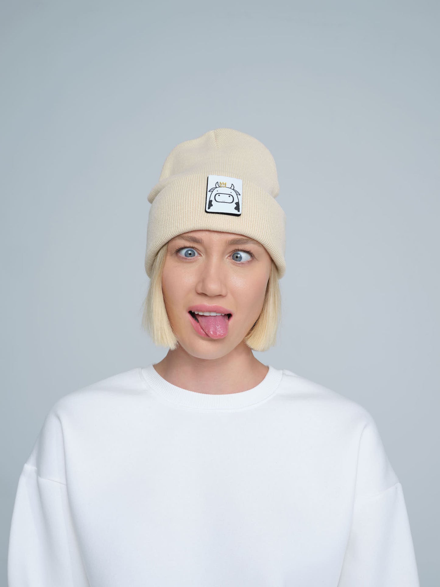 A person wearing a beige beanie hat with a 'cow' design on the cuff, paired with a white t-shirt.
