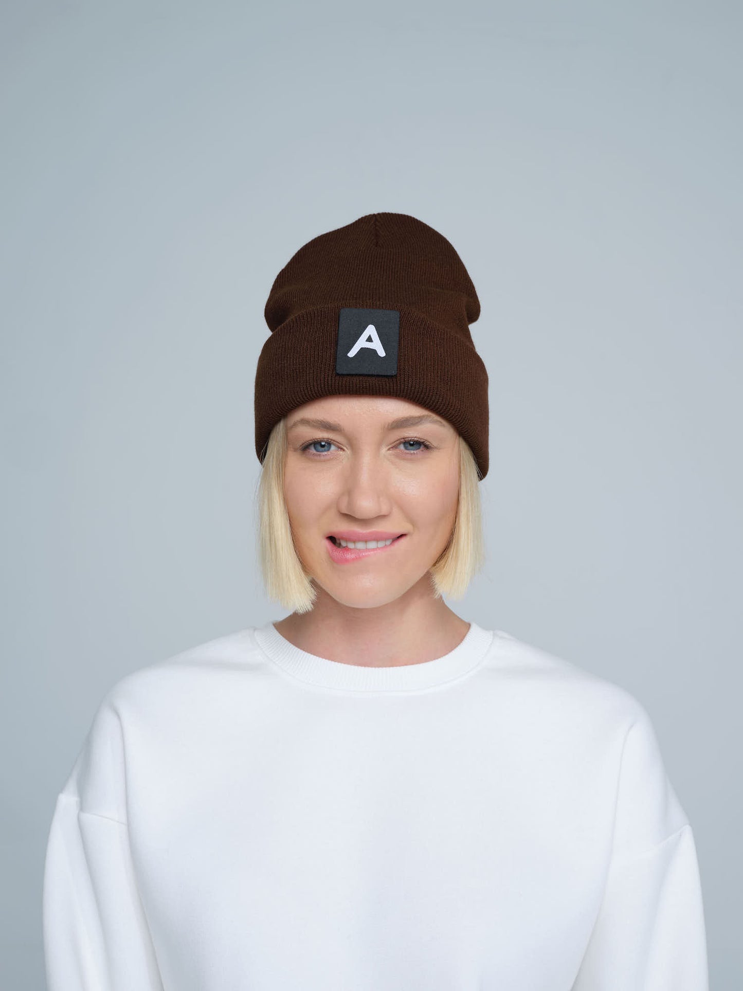 A person wearing a dark brown beanie with a 'A' letter design patch on the front.