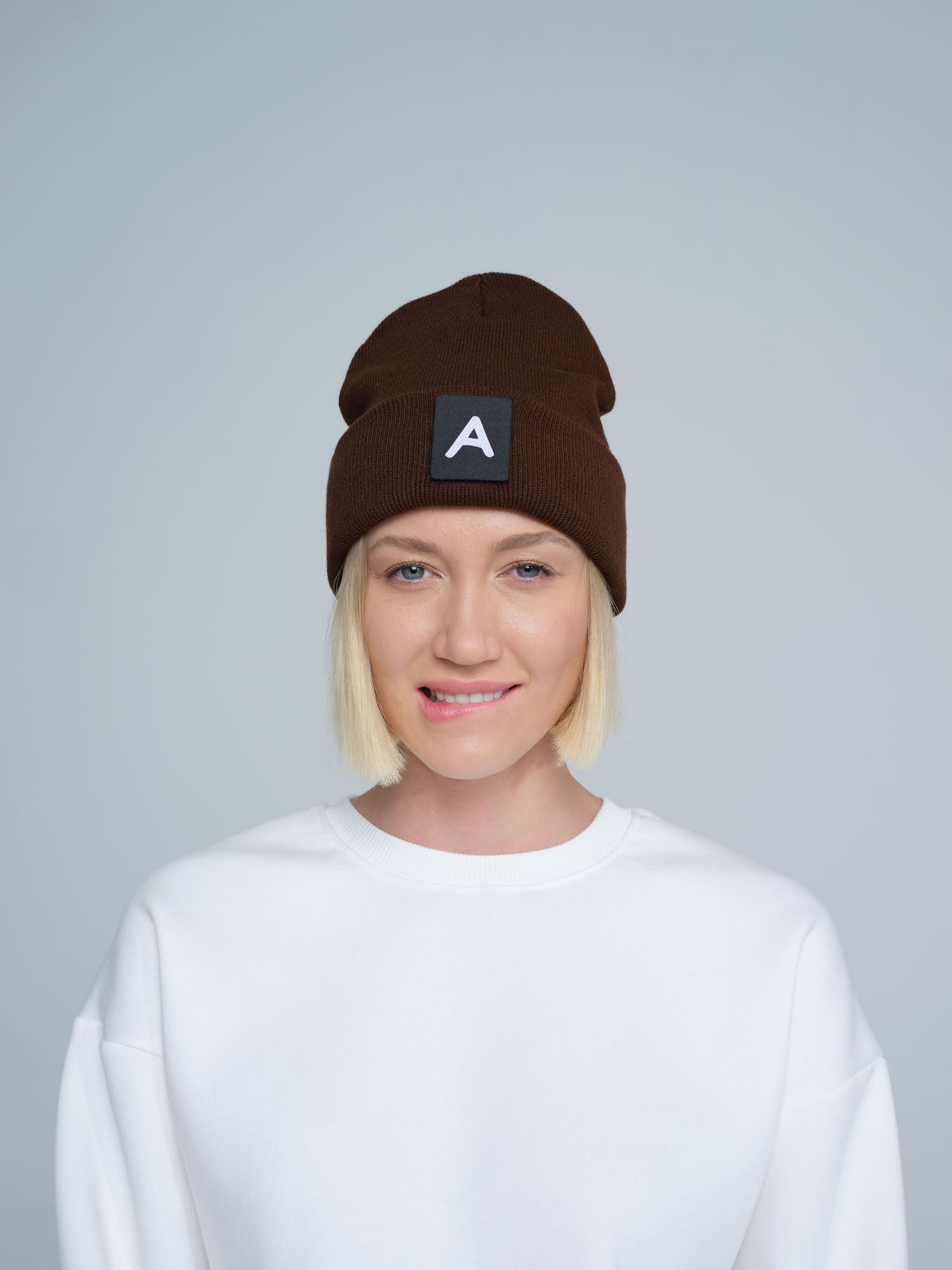 A person wearing a dark brown beanie with a 'A' letter design patch on the front.