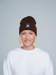 A person wearing a dark brown beanie with a 'A' letter design patch on the front.