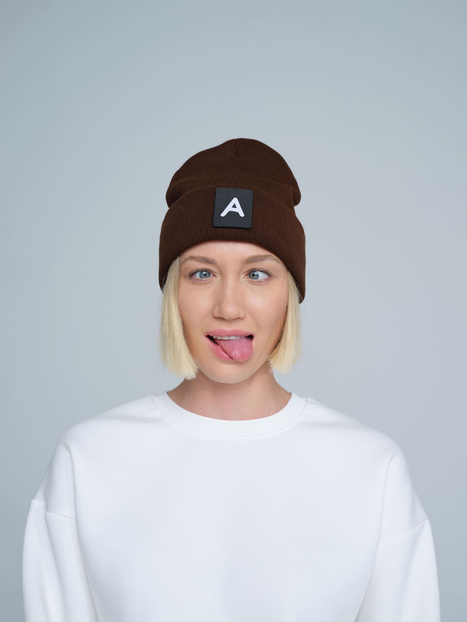 A person wearing a dark brown beanie with a 'A' letter design patch on the front.
