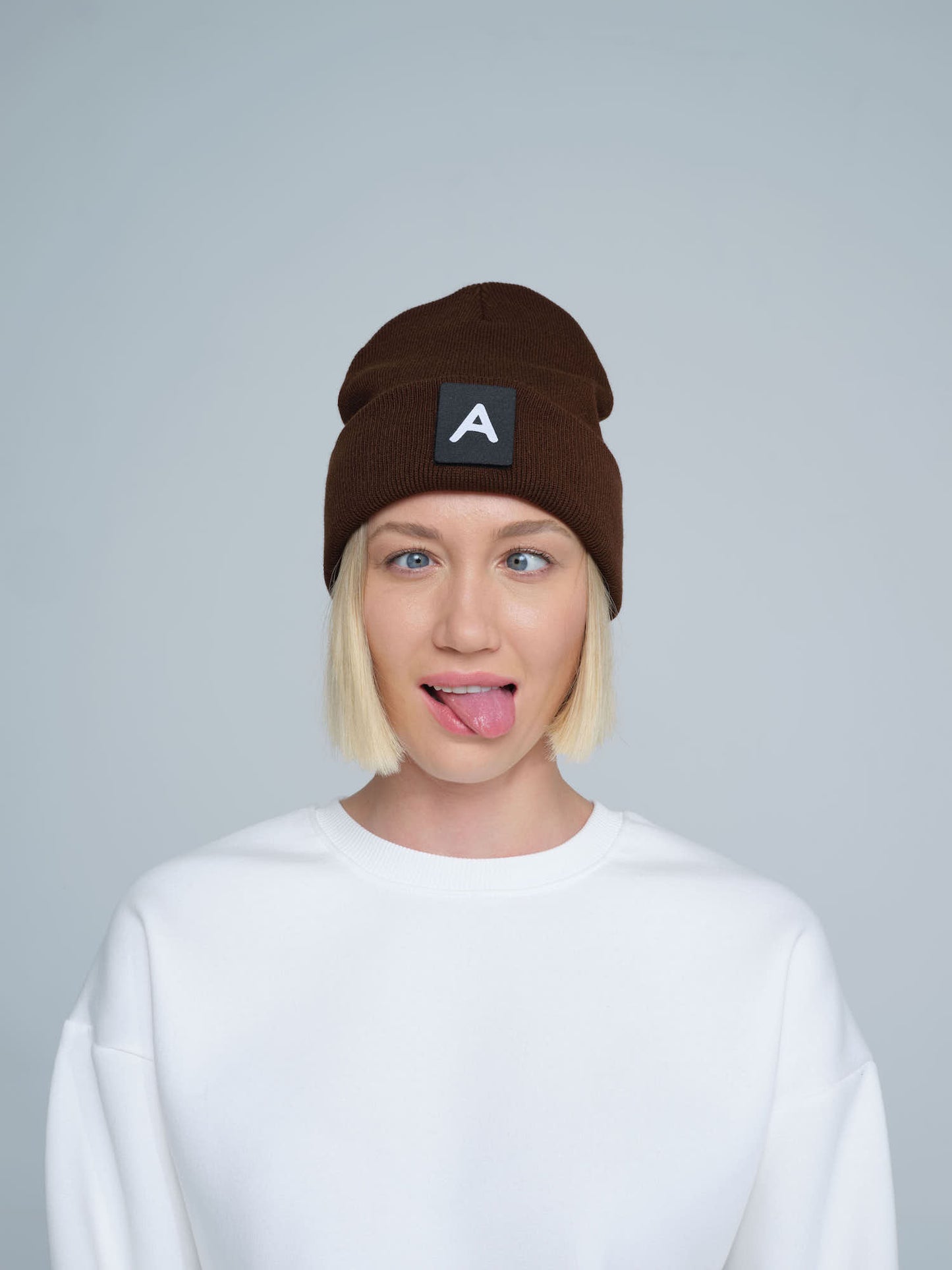 A person wearing a dark brown beanie with a 'A' letter design patch on the front.