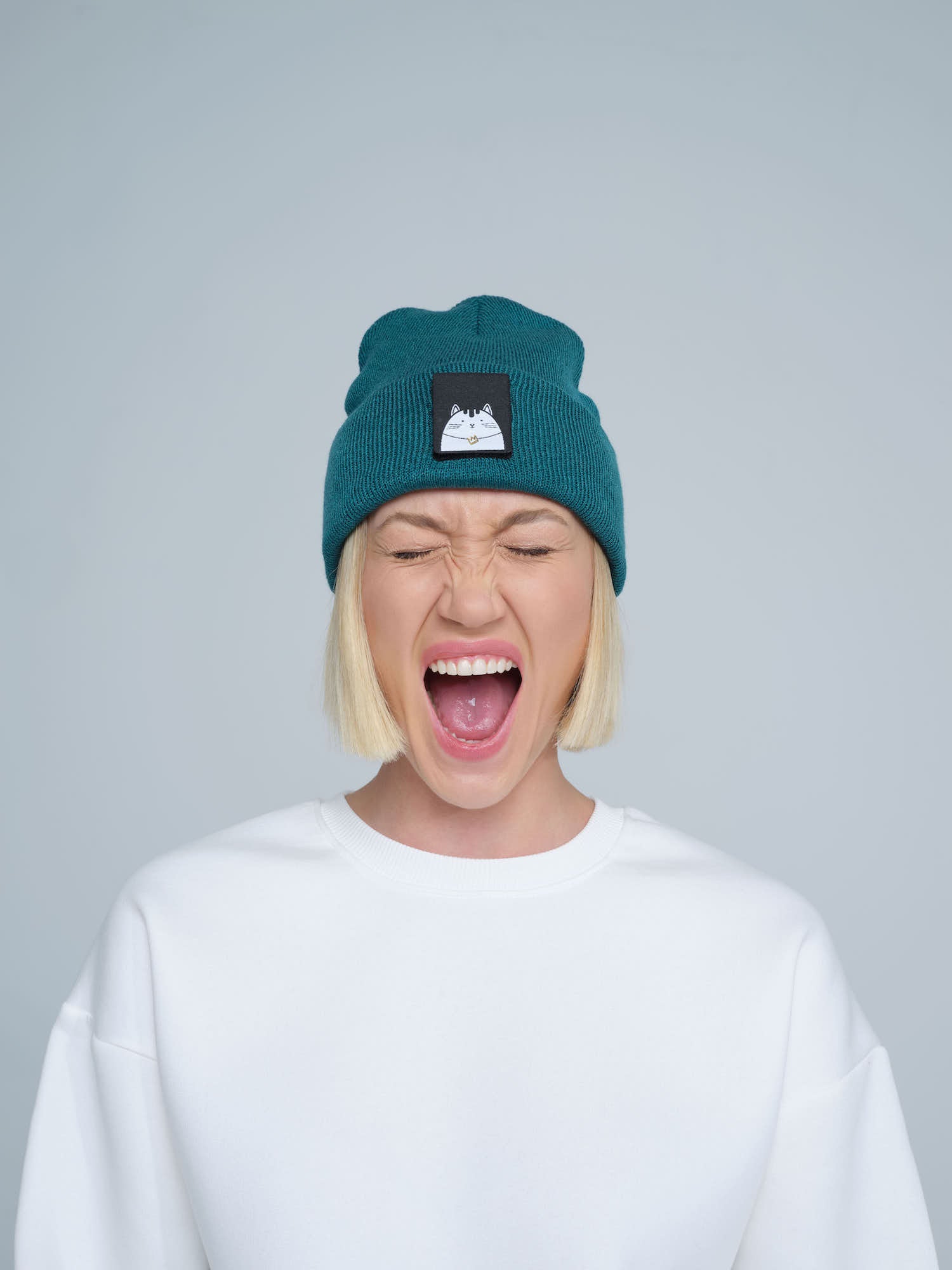A female wearing a emerald beanie hat with a cat design on the cuff, making a scream face. 