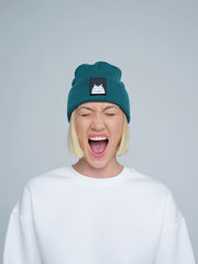 A female wearing a emerald beanie hat with a cat design on the cuff, making a scream face. 