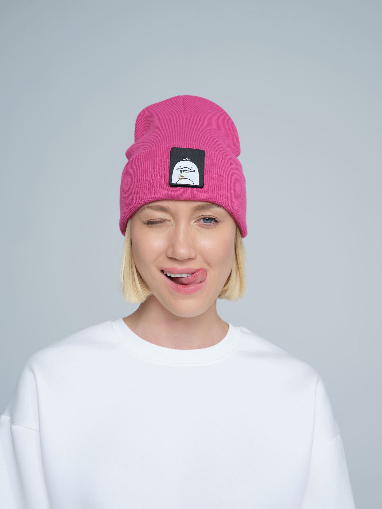 A person wearing a pink beanie with a 'duck' animal design on the fold, part of a white crew neck sweater.