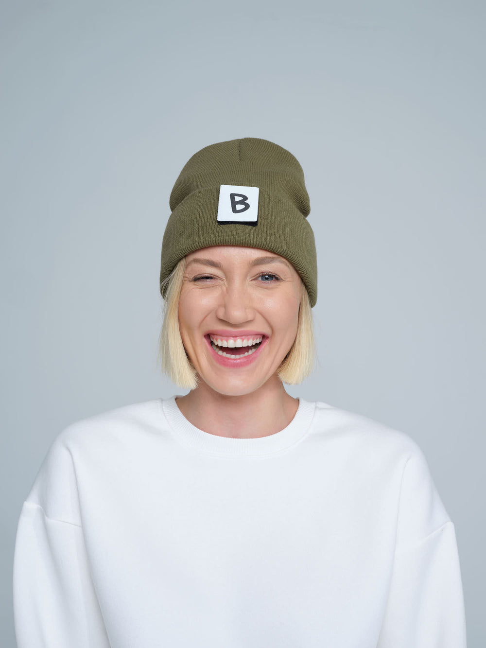 A person wearing an khaki beanie hat with a 'B' design patch on it, smiling against a white background.