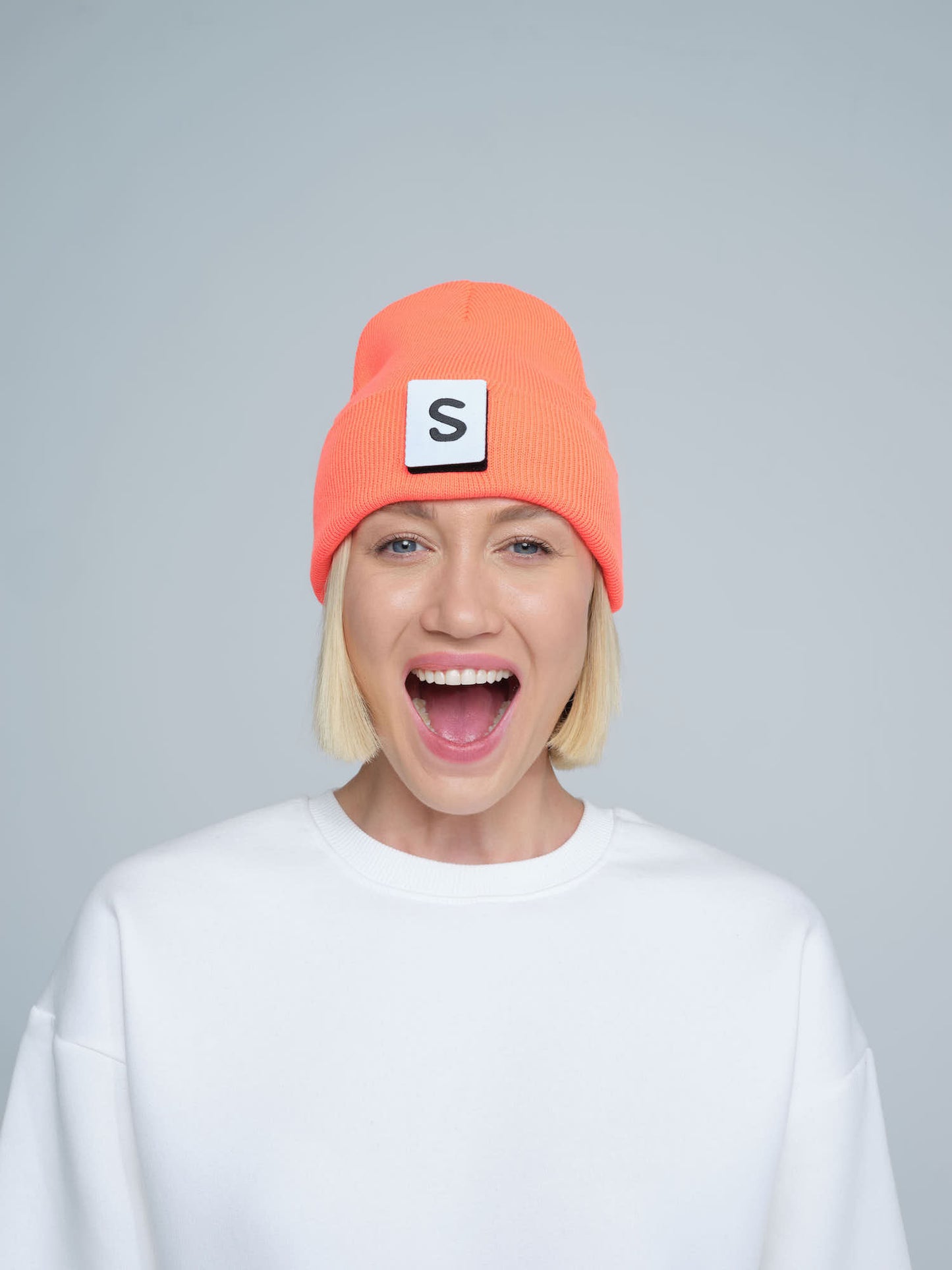 A person wearing a neon orange beanie with a text pattern on the fold, paired with a white t-shirt and smiling.