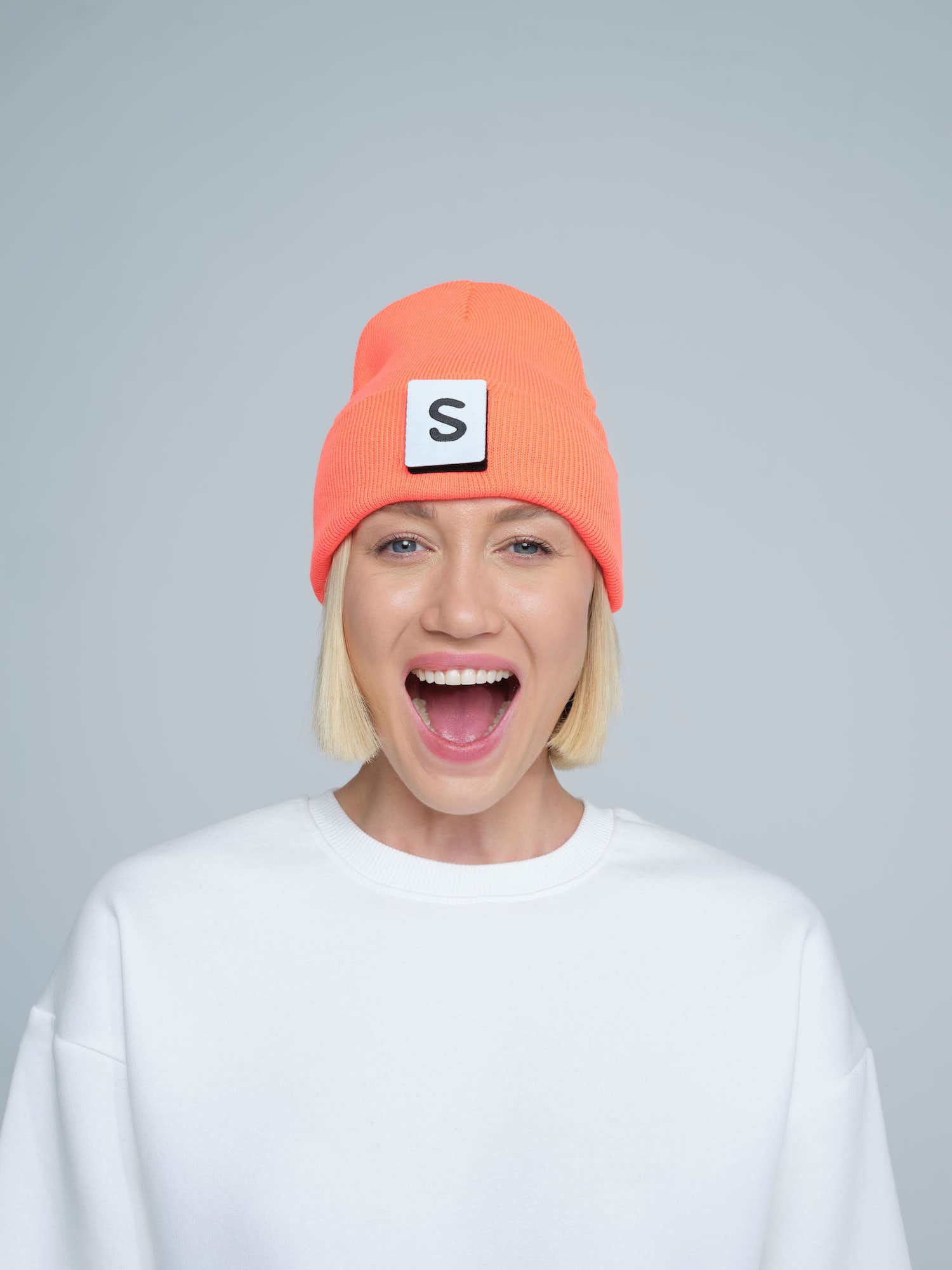 A person wearing a neon orange beanie with a text pattern on the fold, paired with a white t-shirt and smiling.