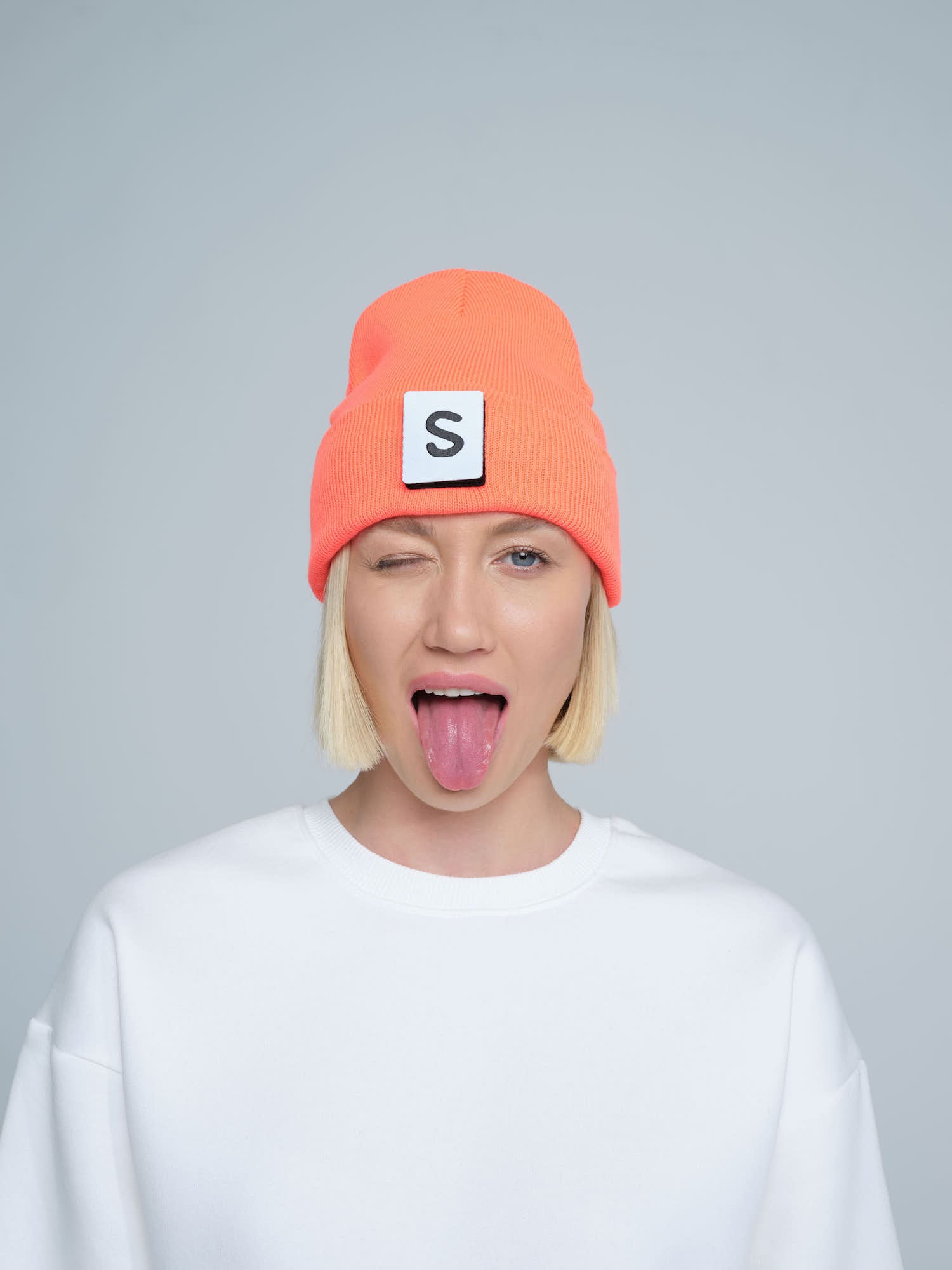 A person wearing a neon orange beanie with a 'S' text pattern on the fold, paired with a white t-shirt.