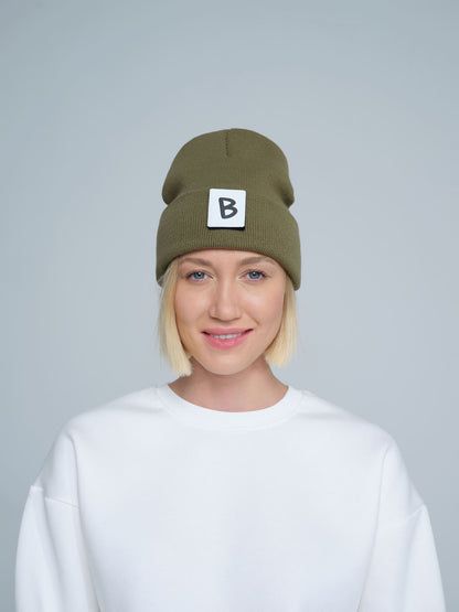 A person wearing an olive green beanie hat with a 'B' design patch on it, smiling against a white background.