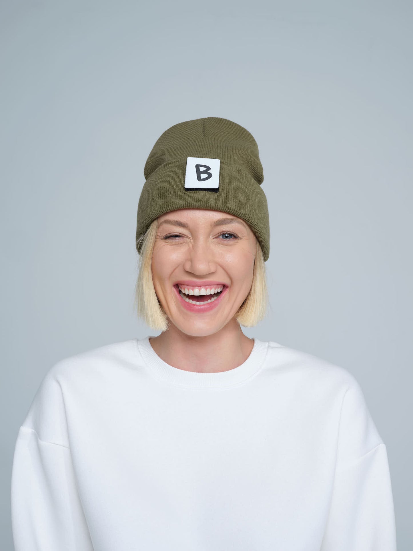 A person wearing an olive green beanie hat with a 'B' design patch on it, smiling against a white background.