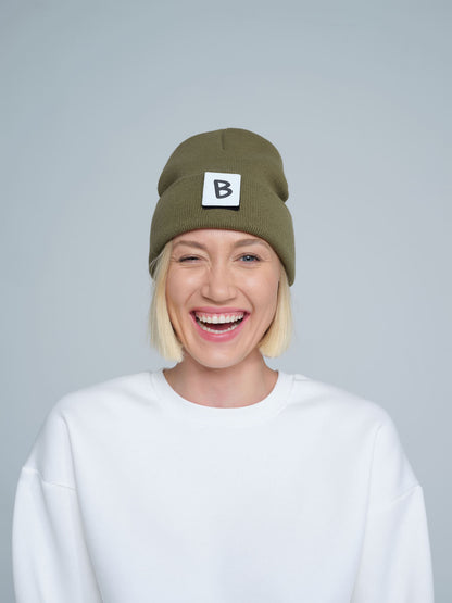 A person wearing an olive green beanie hat with a 'B' design patch on it, smiling against a white background.