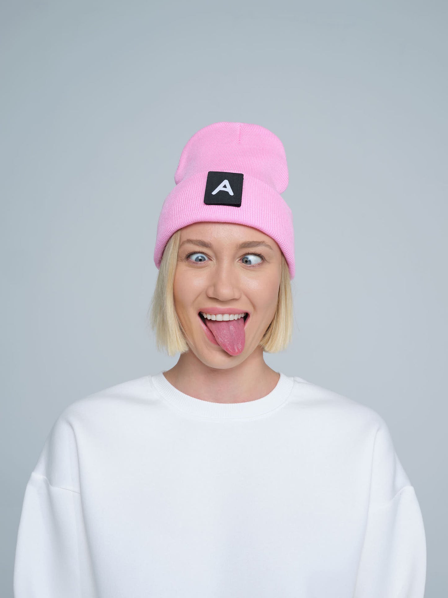 A person wearing a pink beanie with a design on top and the letter 'A' on it, paired with a white crew neck sweater.